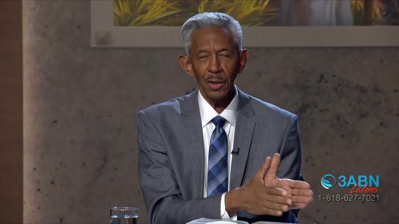 A man in a gray suit gestures with his hands, speaking directly to the camera. The 3ABN Latino logo is visible in the corner, and a glass of water sits on the table in front of him.
