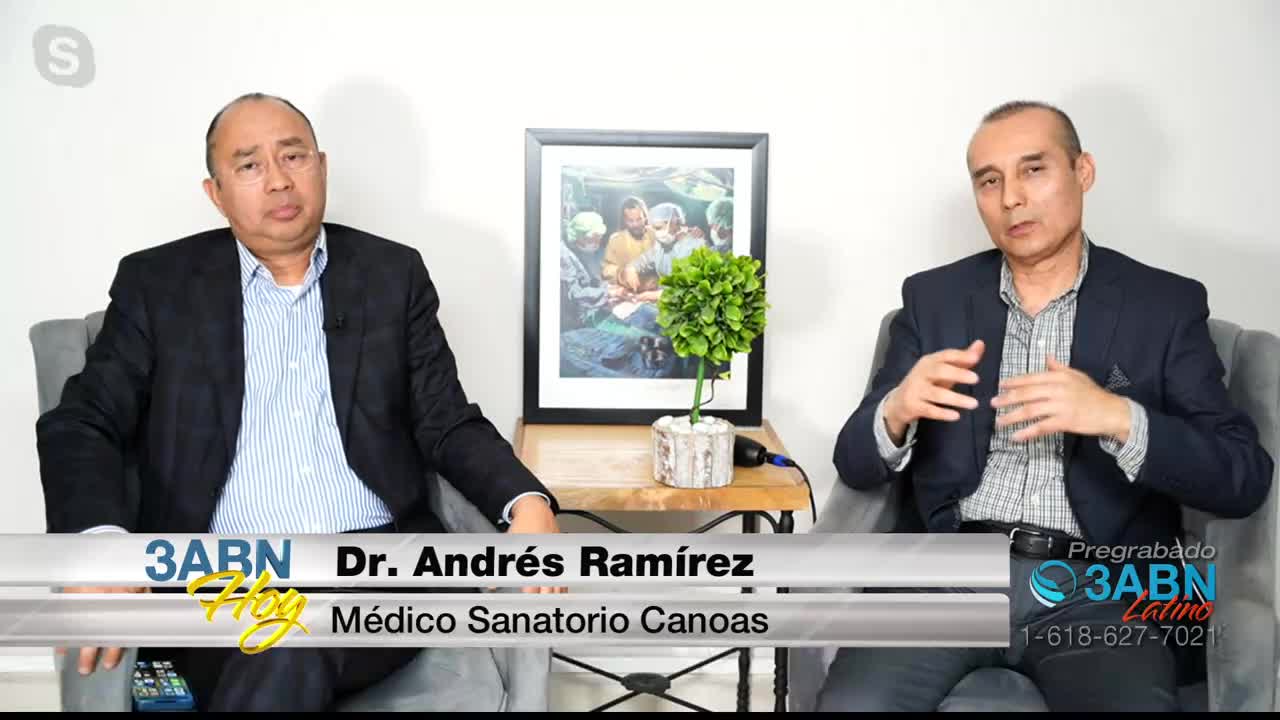 Two men sit in chairs, one on the left and one on the right, facing forward. The man on the right gestures with his hands as he speaks. A framed picture hangs on the wall behind them.
