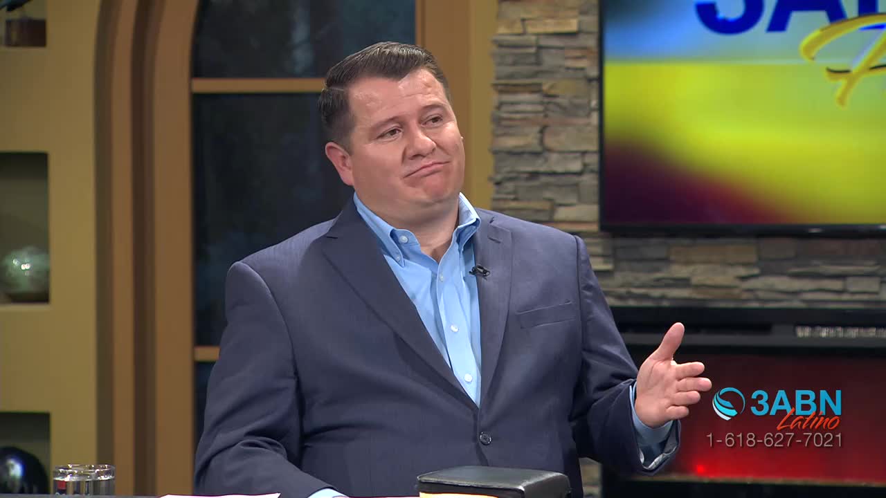A man in a blue suit and light blue shirt gestures with his right hand. Behind him, a screen displays the 3ABN Latino logo.