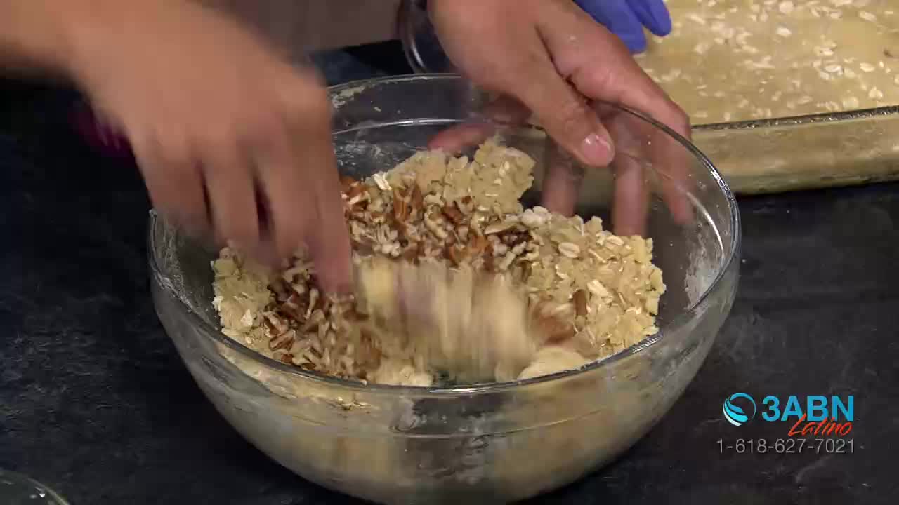 Hands mix chopped pecans and oats into a buttery crumble topping in a clear glass bowl. A baking dish filled with a similar mixture sits in the background.
