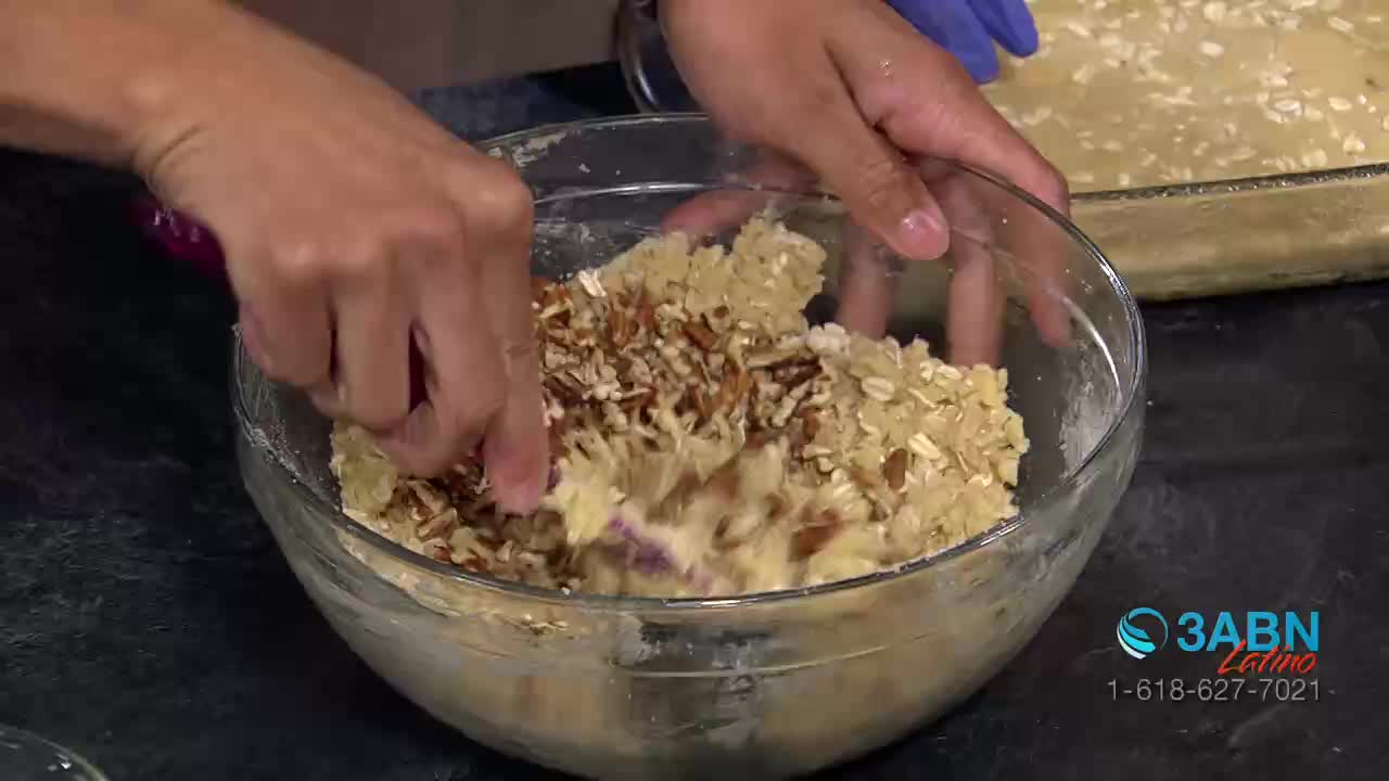 Hands mix chopped pecans into a bowl of batter. A baking dish filled with a similar mixture sits nearby.