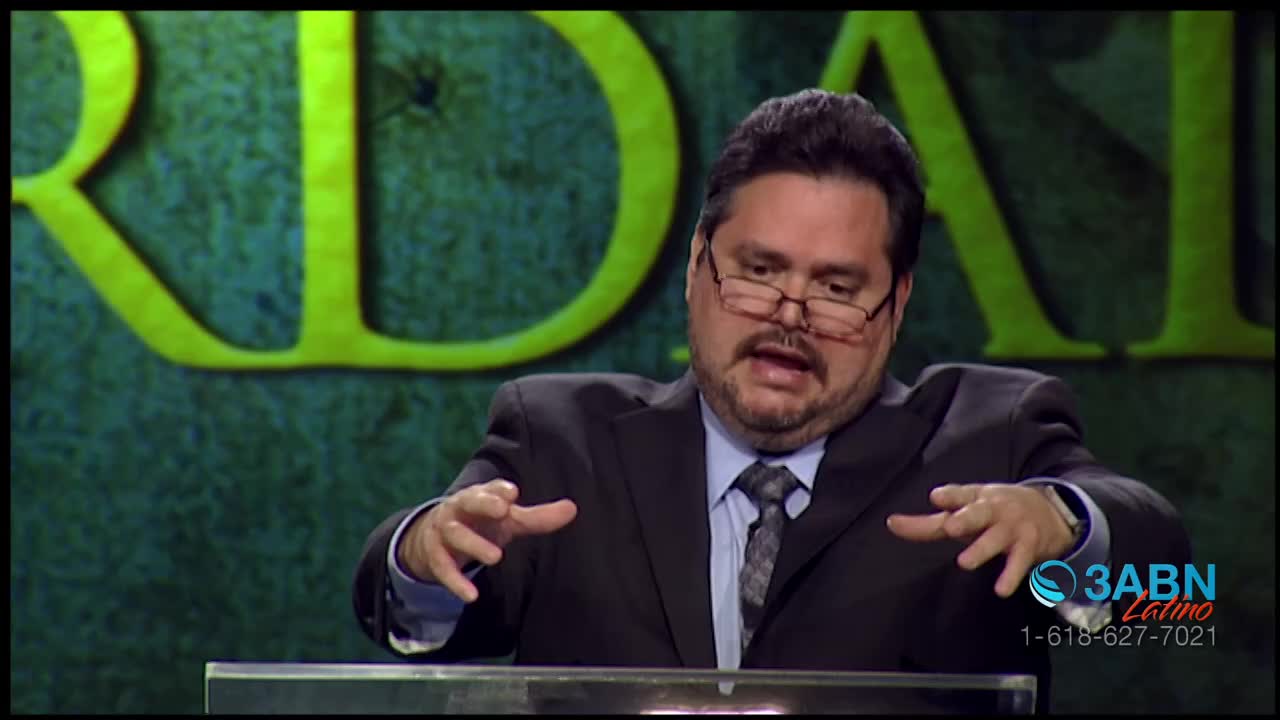 A man in a suit gestures with both hands while speaking. He's on a stage with large yellow letters behind him, and a 3ABN Latino logo is visible.