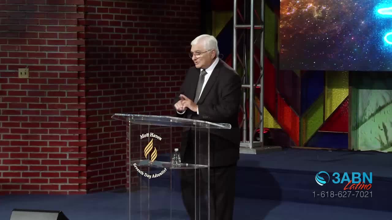 A man in a suit stands at a clear podium, gesturing with his hands as he speaks. Behind him, a large screen displays a cosmic scene, and the 3ABN Latino logo is visible in the lower right corner.