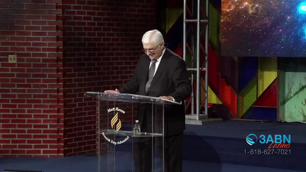 A man in a suit stands at a clear podium, looking down as he speaks. Behind him, a large screen displays a vibrant cosmic scene, and the 3ABN Latino logo is visible on the right.