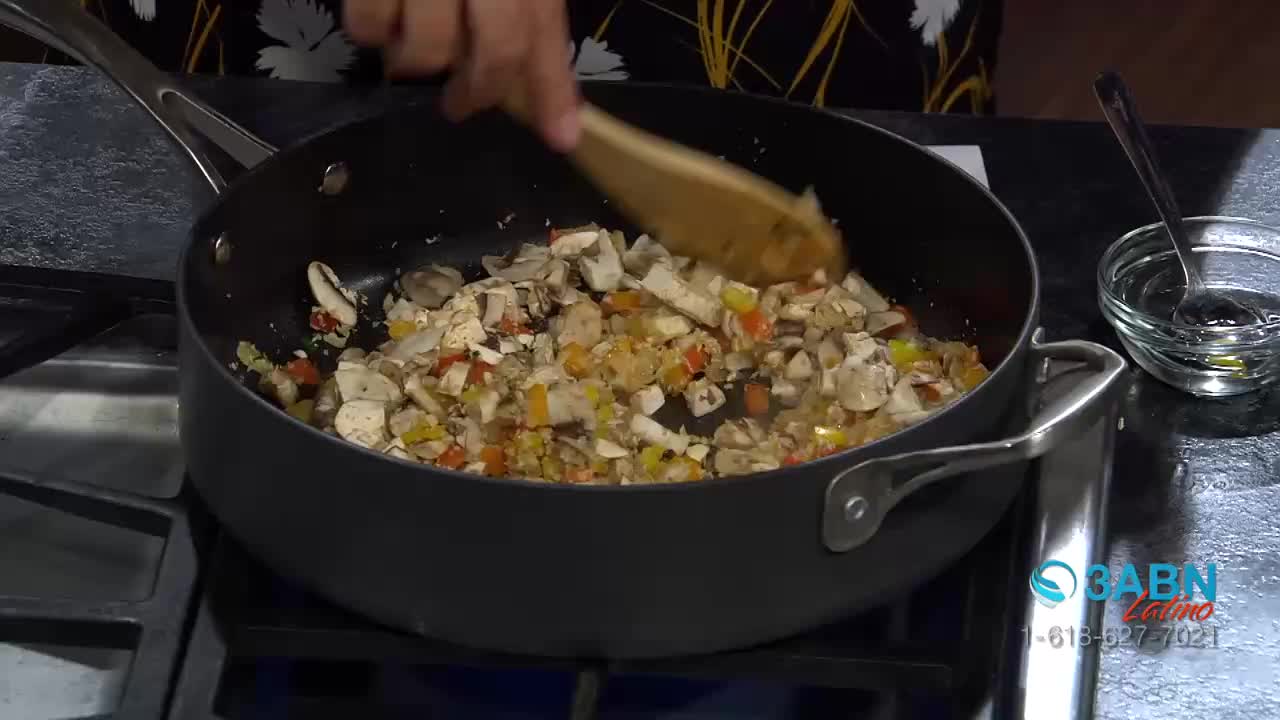 Chopped mushrooms and peppers tumble in a hot pan as a wooden spoon stirs them. A small bowl of dark liquid sits to the side, ready for use.