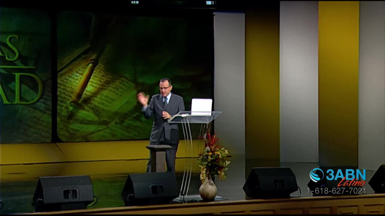 A man in a suit stands at a podium, gesturing with his right hand. Behind him, a large screen displays an open book with a magnifying glass. The 3ABN Latino logo appears in the lower right corner.