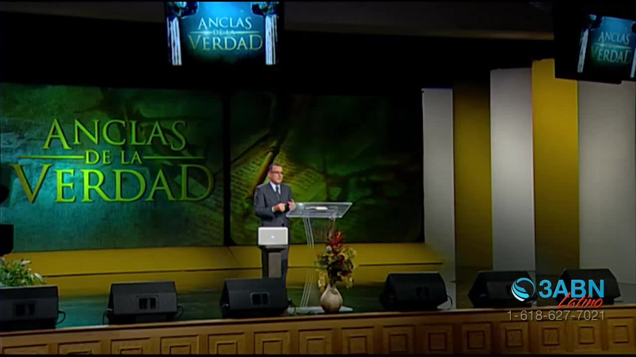 A man in a suit stands behind a clear podium, speaking to an unseen audience. The stage is set with a large green backdrop featuring the words "Anclas de la Verdad" in yellow lettering.