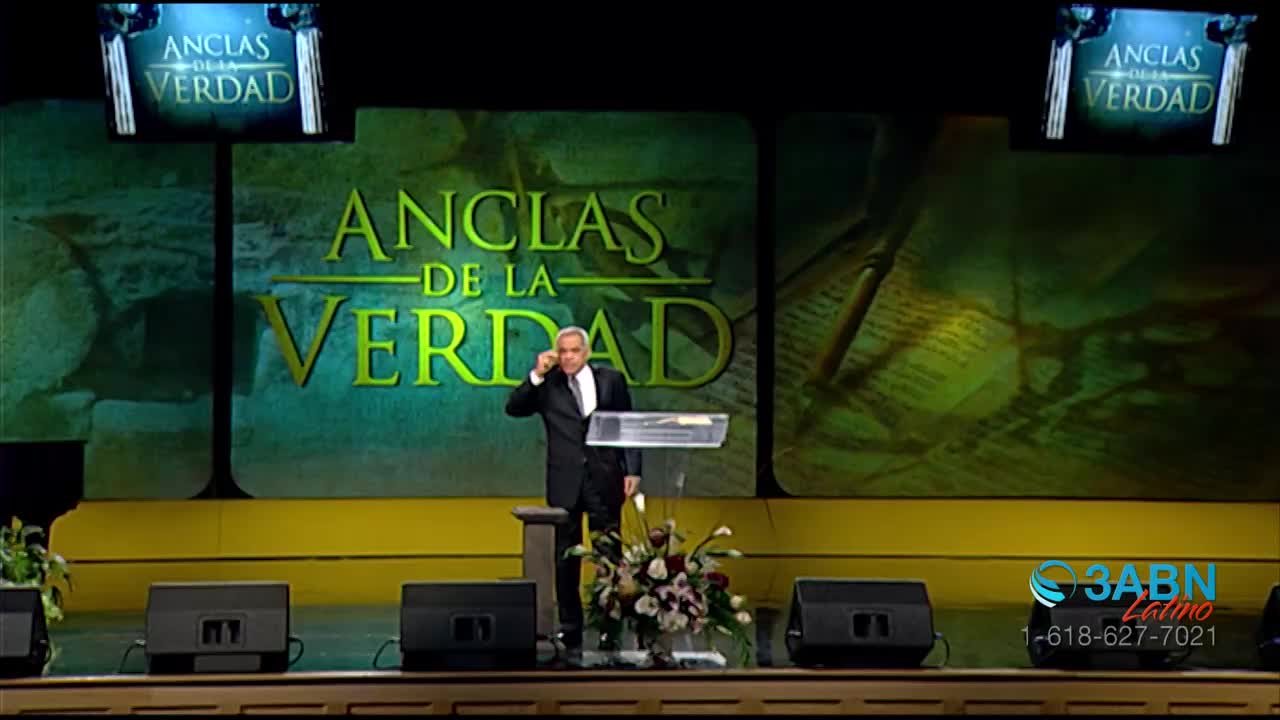 A man in a suit stands at a podium on a stage, gesturing with his hand as if making a point. Behind him, large green screens display the words "ANCLAS DE LA VERDAD" in bold lettering.