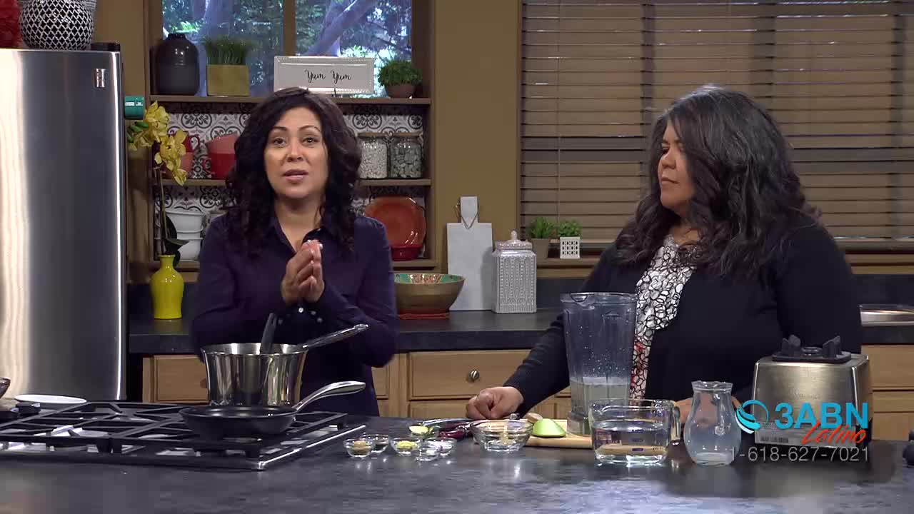 Two women stand in a kitchen, one speaking with her hands clasped. A stainless steel pot sits on a stovetop in front of her, and a blender is visible behind the other woman. The 3ABN Latino logo appears on a device in the foreground.