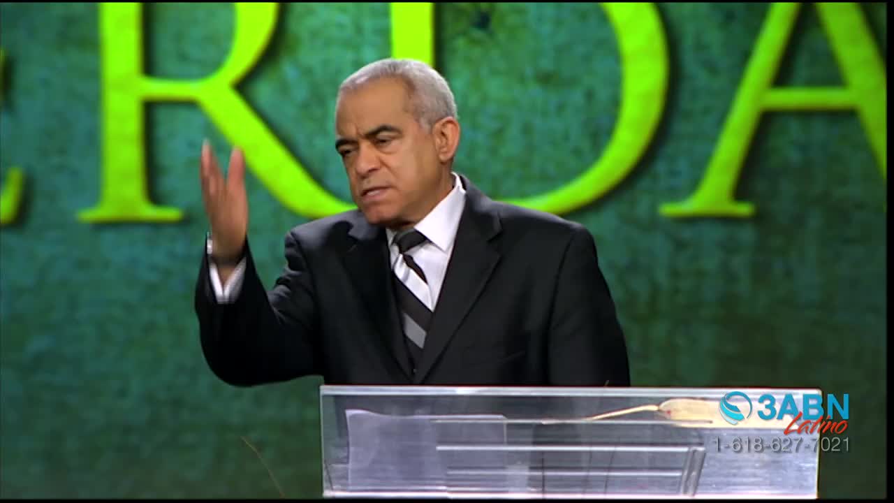 A man in a suit gestures with his right hand while speaking from behind a clear podium. A logo for 3ABN Latino appears on the lower right.