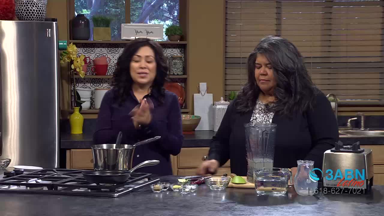 Two women stand in a kitchen, likely preparing a recipe for 3ABN Latino. One woman claps her hands together while standing next to a pot on the stove, and the other woman is positioned by a blender.