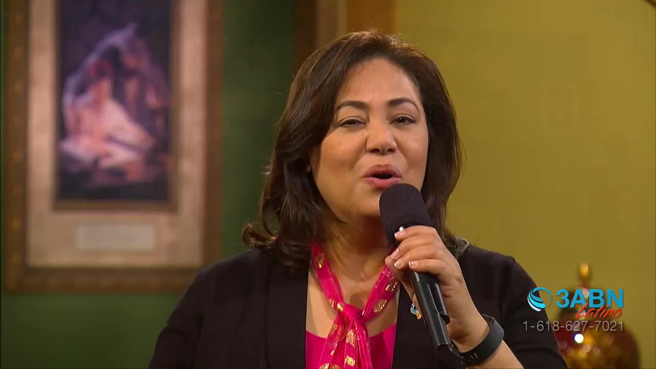 A woman speaks into a microphone, her expression animated. Behind her, a framed painting hangs on the wall.