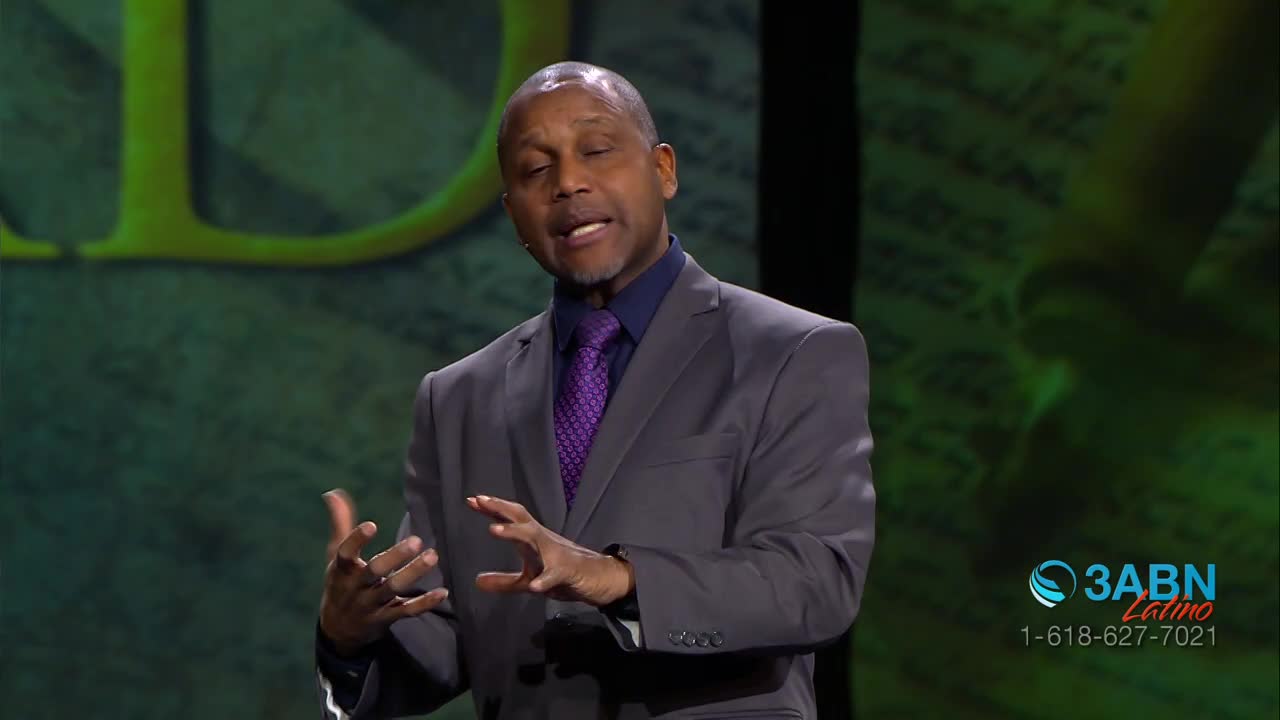 A man in a gray suit and purple tie speaks with his hands, gesturing animatedly. Behind him, a green backdrop displays a large, stylized letter and what appears to be text. A 3ABN Latino logo and phone number are visible in the lower right corner.