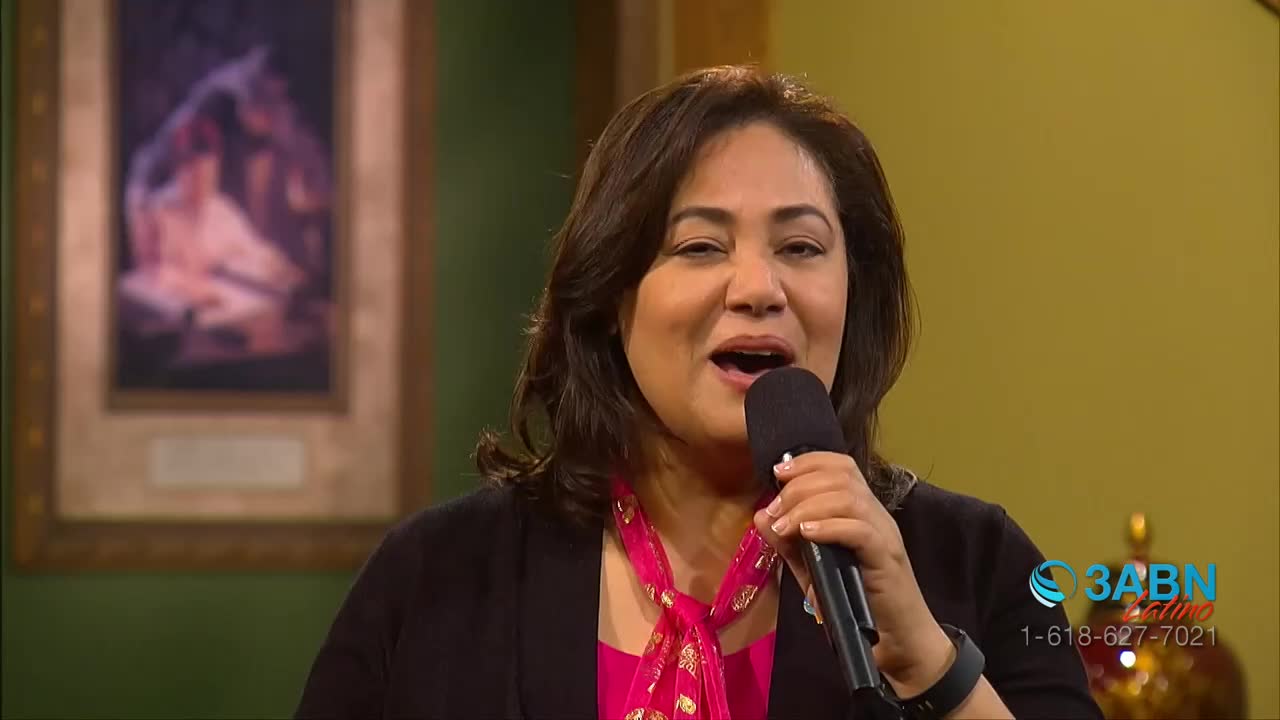 A woman with dark hair and a bright pink scarf is speaking into a microphone. Behind her, a framed painting hangs on a green wall.