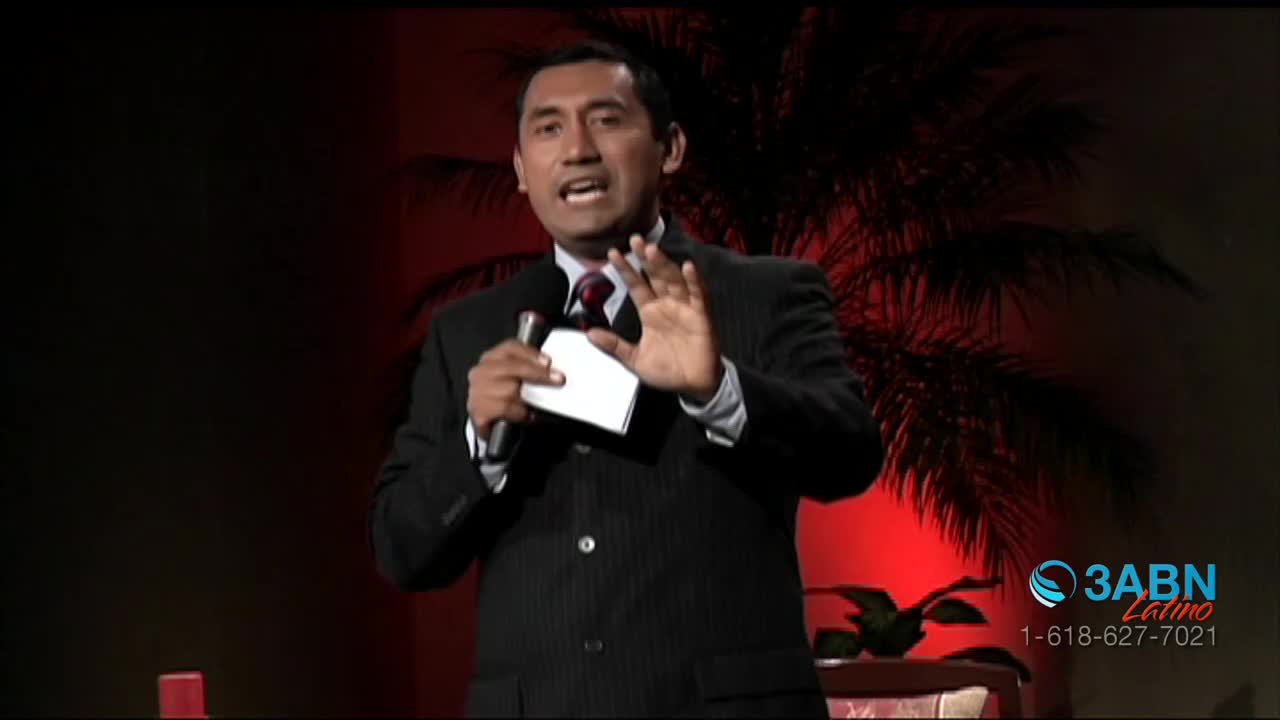 A man in a pinstripe suit stands at a podium, holding a microphone and a small white card. He gestures with his open hand as he speaks, his expression earnest. Behind him, a palm frond casts a shadow against a red-lit backdrop.