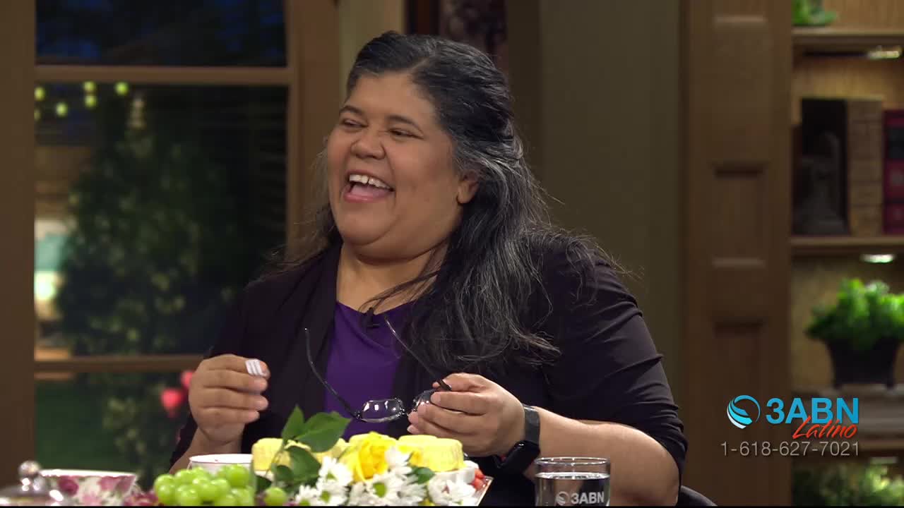 A woman laughs heartily, her head tilted back. She holds a pair of glasses in her hands, gesturing as if sharing a joke on the 3ABN Latino program.