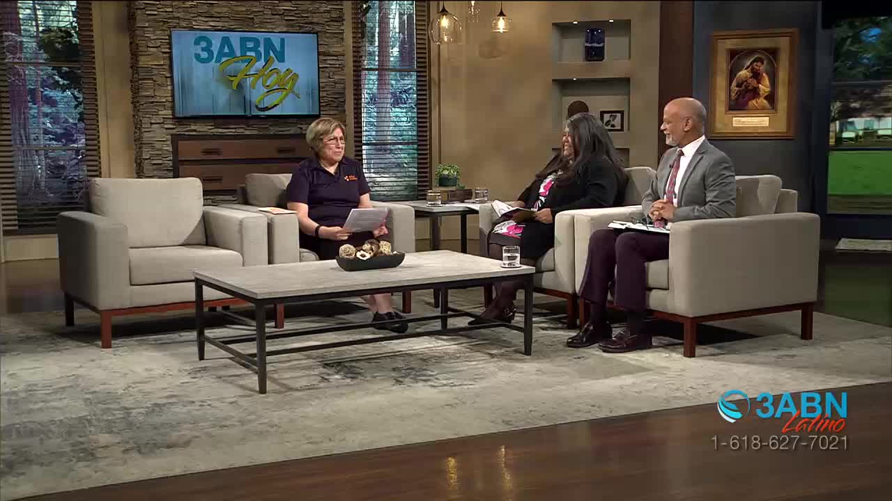 The woman in the purple shirt holds papers, looking toward the camera. A man in a suit sits across from her, his hands clasped.