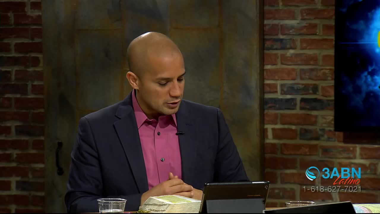A man in a dark suit and pink shirt speaks, looking down at a tablet and a Bible. The 3ABN Latino logo appears on a screen behind him.