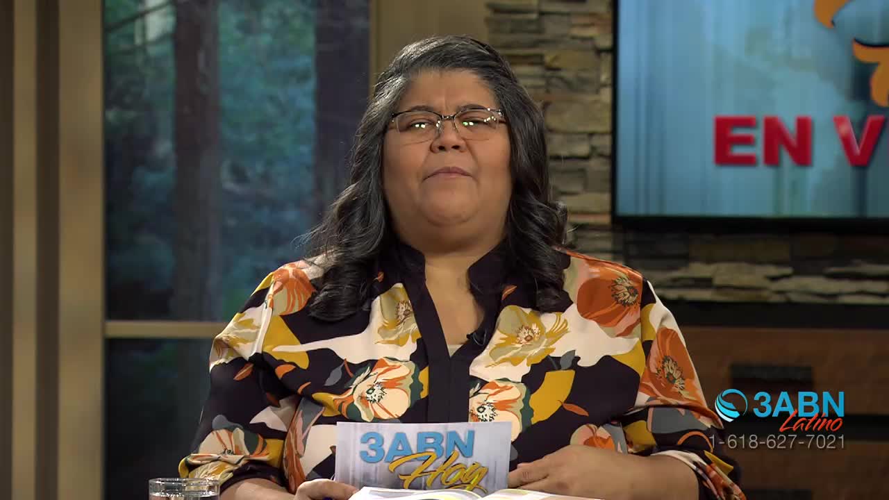 A woman with dark, wavy hair and glasses looks directly ahead. She wears a colorful blouse with a floral pattern, and a sign with the 3ABN Latino logo rests on a book in front of her.