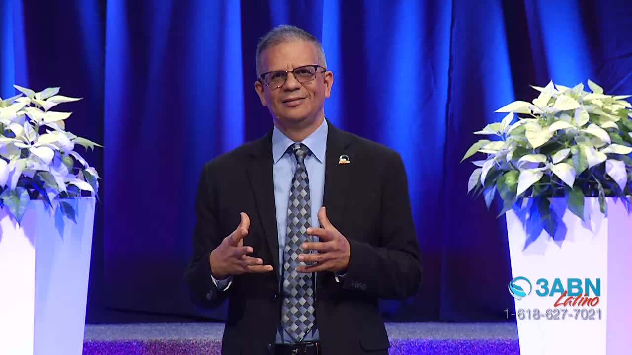 A man in a suit speaks with his hands gesturing, standing between two large white planters. The 3ABN Latino logo appears on the right side of the stage.