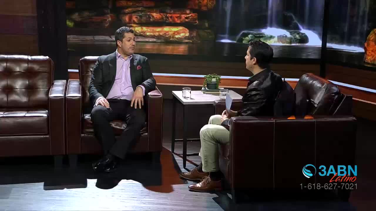 Two men sit across from each other in plush chairs on a studio set. A waterfall backdrop suggests a serene setting for their conversation.