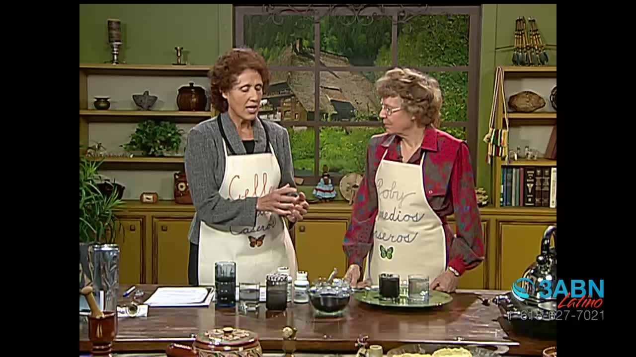 Two women stand behind a counter, each wearing an apron. One woman gestures with her hands as she speaks, while the other listens intently. On the counter in front of them are several glasses and small containers.
