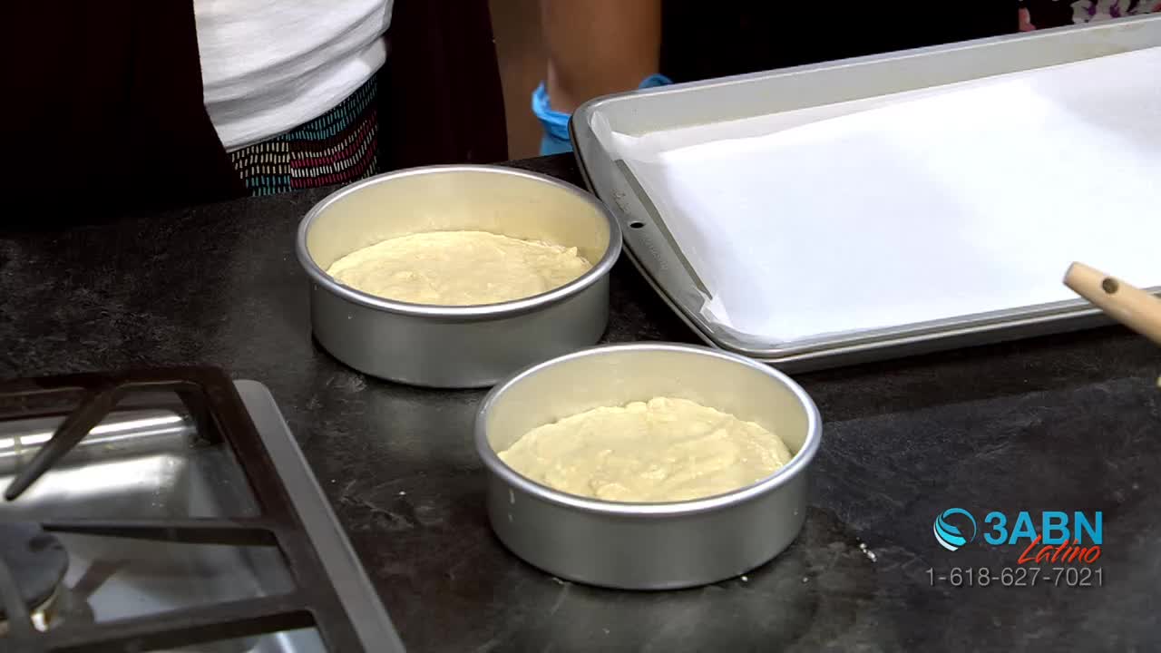 Two round cake pans sit on a dark countertop, each filled with a pale yellow batter. Beside them, a large rectangular baking sheet lined with white parchment paper awaits its turn.