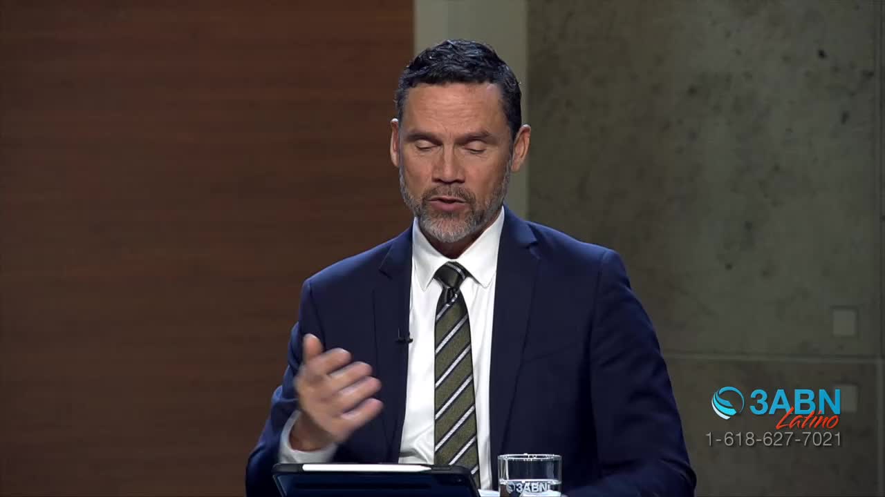 A man in a suit speaks with his hands, gesturing towards the left. A glass of water sits on the table beside him, and the 3ABN Latino logo appears in the corner.