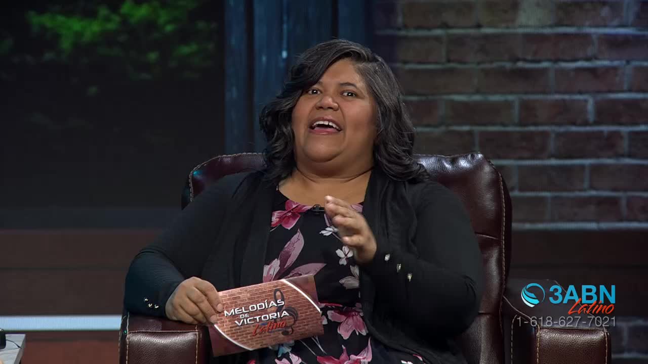 A woman with dark, wavy hair speaks animatedly, holding a card that reads "Melodías de Victoria." The 3ABN Latino logo and a phone number are visible on the right side of the screen.