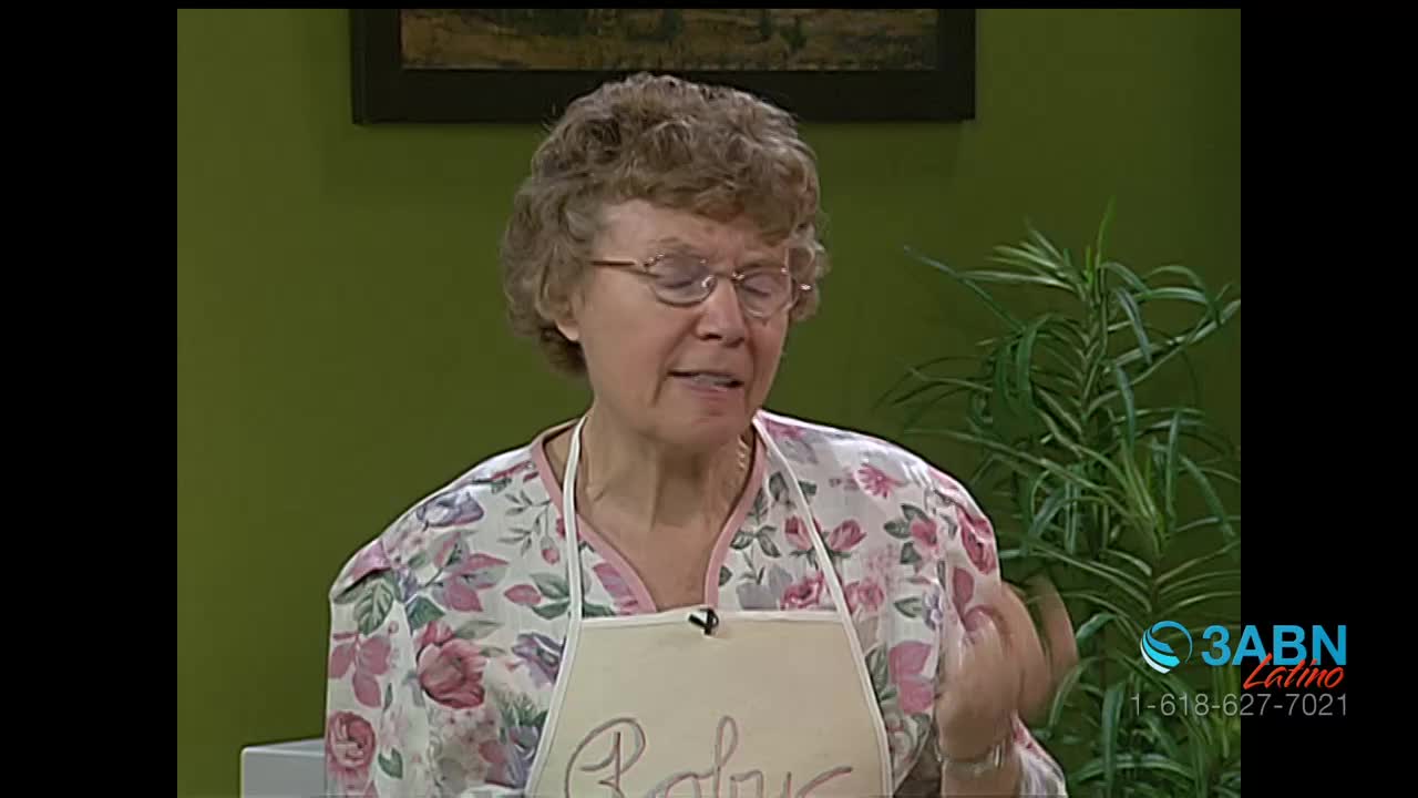A woman with permed hair and glasses speaks animatedly, her hands gesturing as she talks. She wears a floral blouse and a white apron that reads "Baby."