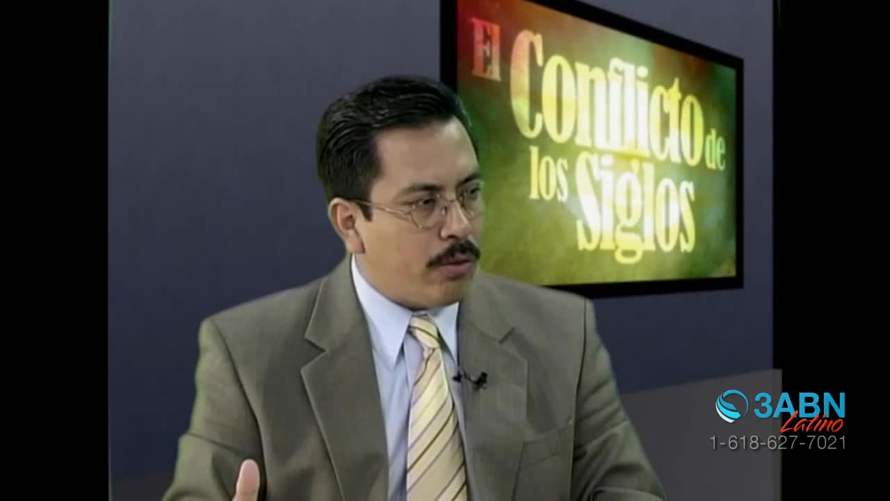 A man in a suit speaks, gesturing with his hand. Behind him, a screen displays "El Conflicto de los Siglos" in bright yellow lettering.