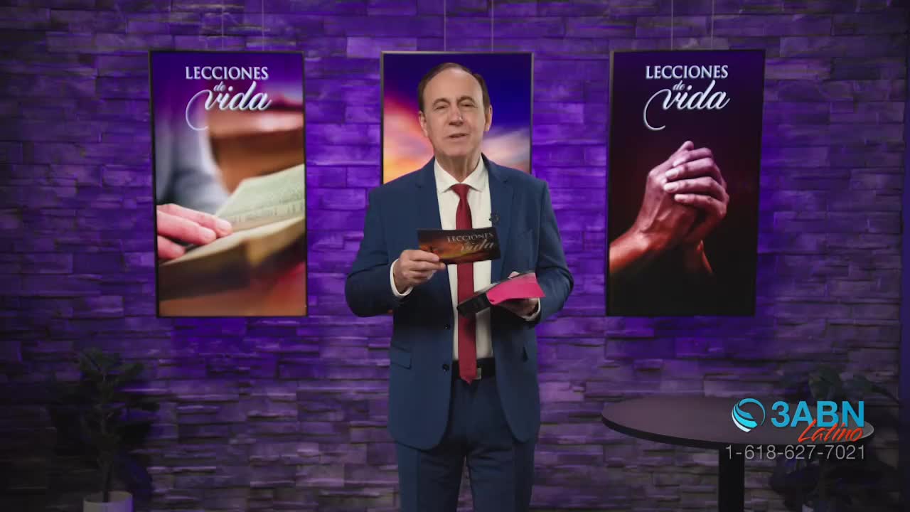 A man in a suit stands holding a small card and a pink booklet. Behind him, two large screens display the words "Lecciones de Vida" over images of hands reading a book and hands clasped in prayer. The 3ABN Latino logo appears in the lower right corner.