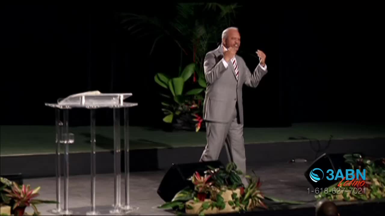A man in a gray suit speaks from a stage, gesturing with his hands. Tropical plants surround the stage, and a logo for 3ABN Latino appears in the corner.