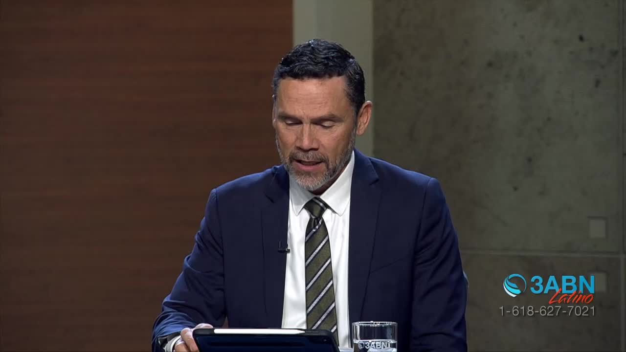 A man in a dark suit and striped tie speaks, looking down at a tablet. A glass of water sits to his right, next to a logo for 3ABN Latino.