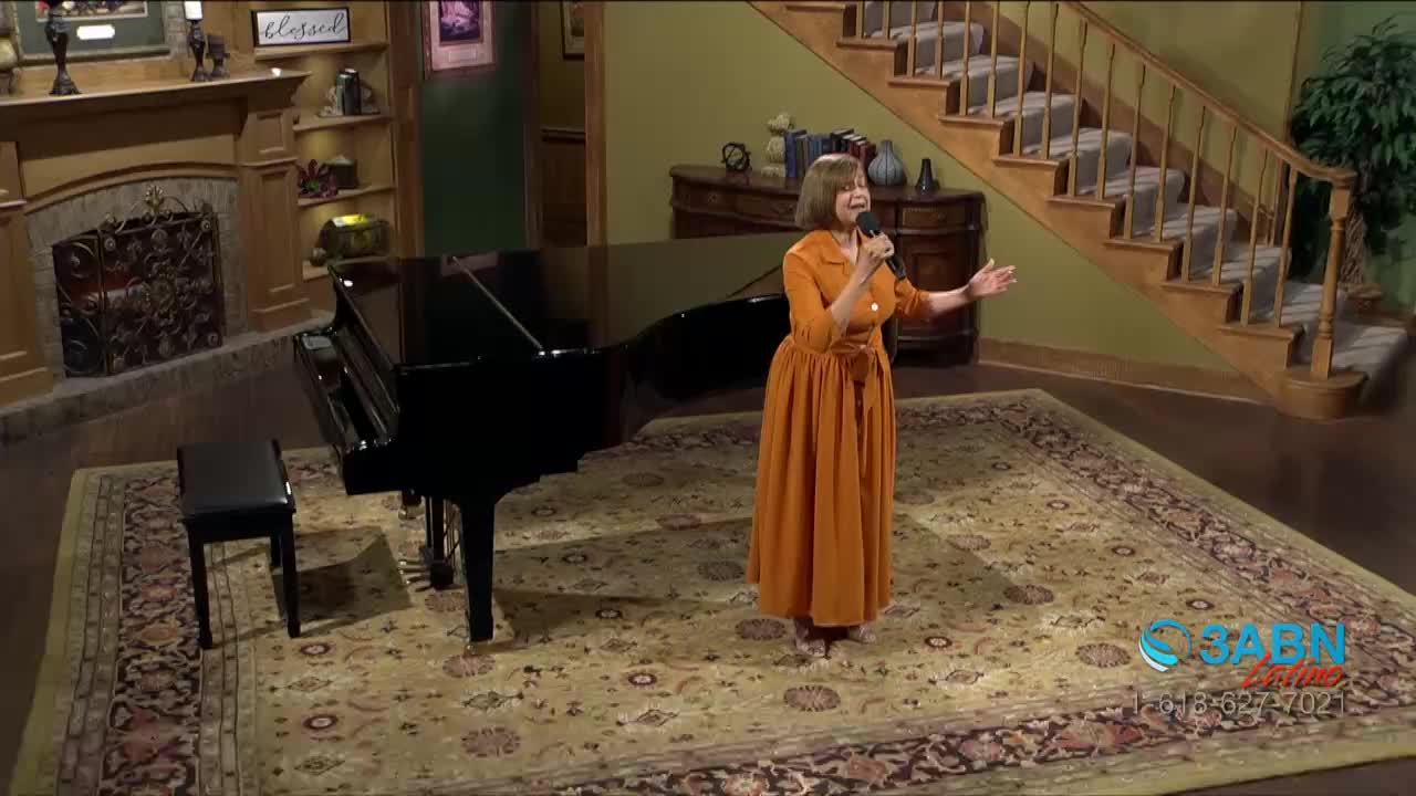 A woman in an orange dress is singing into a microphone, her arms outstretched. A grand piano sits to her left, its polished surface reflecting the light.