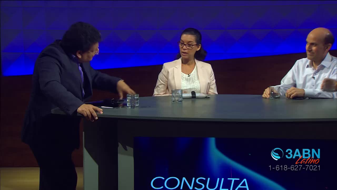 A man in a dark suit leans over a table, his hand reaching for a tablet. Across from him, a woman in a light jacket and a man in a white shirt sit engaged in conversation. The 3ABN Latino logo is visible on a screen below the table.
