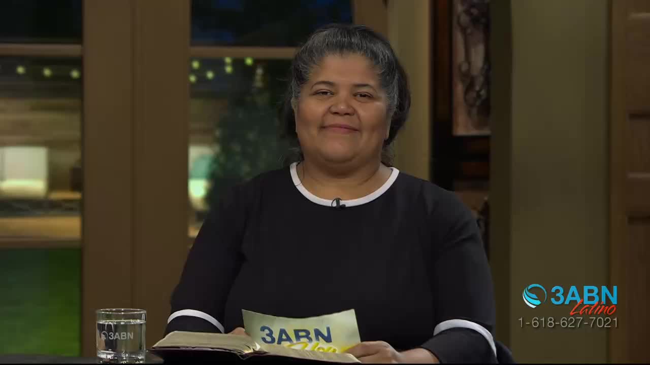 A woman in a black shirt with white trim smiles directly at me. She holds a yellow card with "3ABN Latino" printed on it.