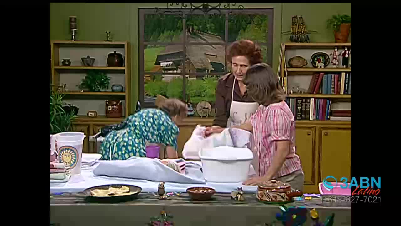 A woman in a green dress leans over a table, her back to me, while two other women stand nearby. One woman, wearing an apron, looks down at a white basin on the table.