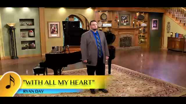 A man in a suit stands center stage, holding a microphone in a studio setting. Behind him, a grand piano sits on a patterned rug, and the set is decorated with a fireplace and artwork.
