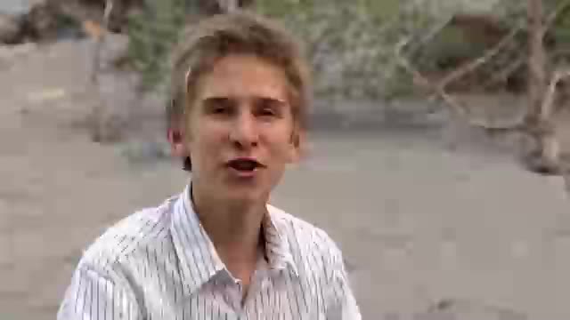 A man in a striped shirt is speaking directly into the camera, his mouth moving as he talks. The background is an out-of-focus, sandy area with some sparse vegetation.

