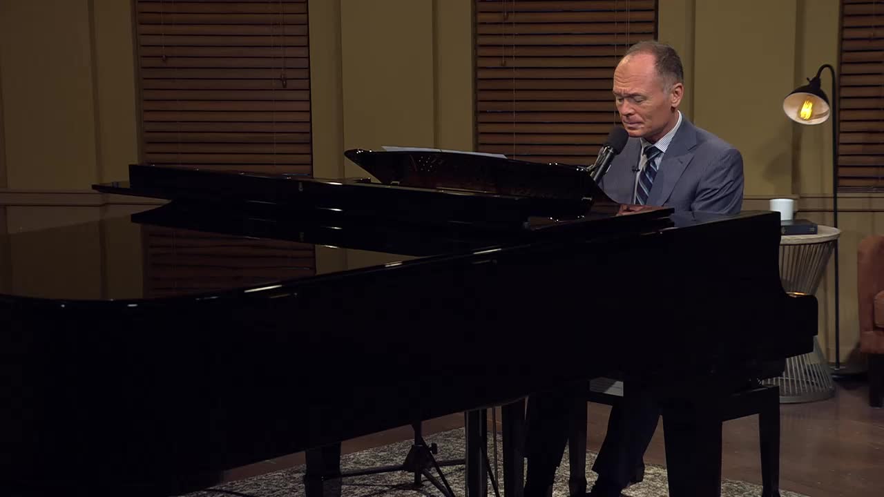 A man in a suit sits at a grand piano, his fingers poised over the keys. He's looking down at the music, ready to play.
