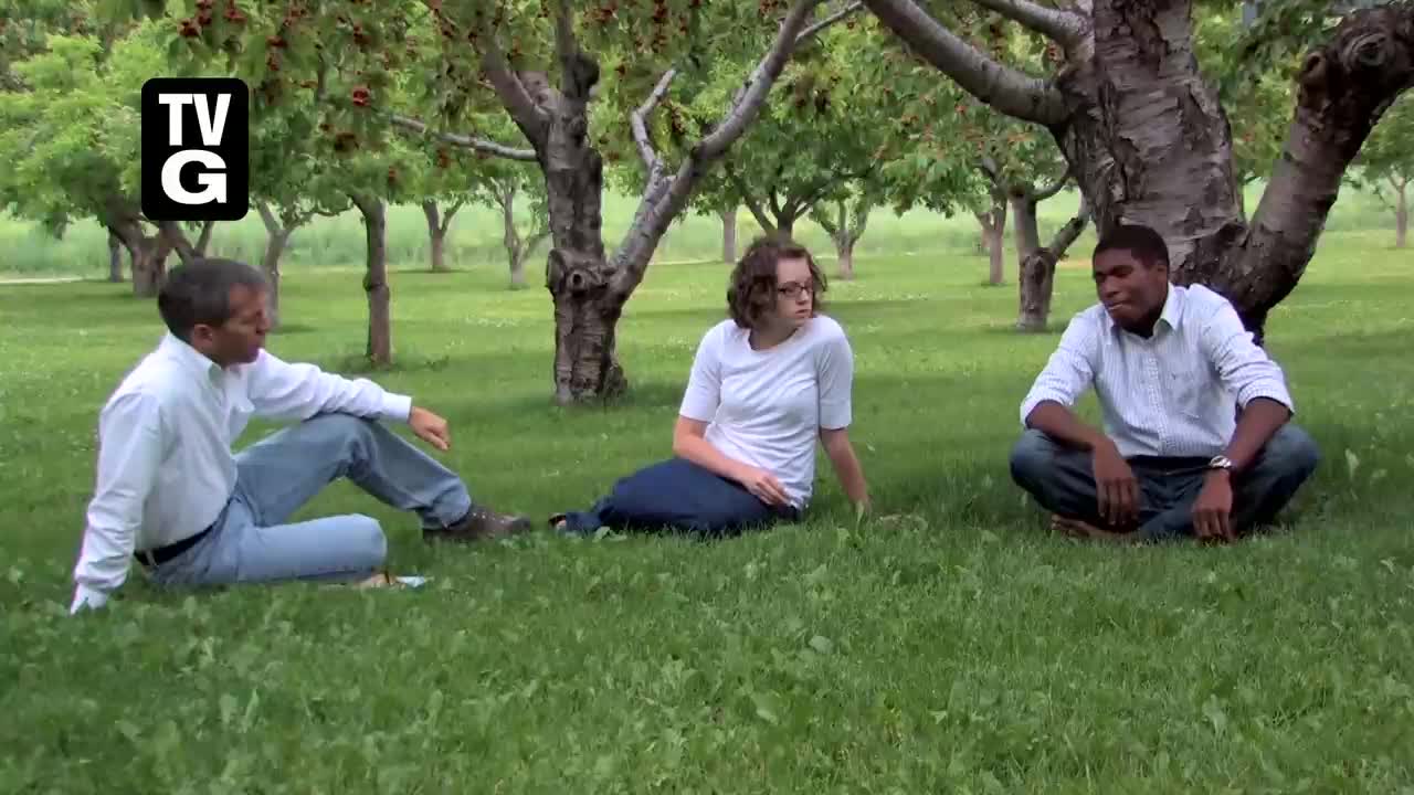 Three people are sitting on the grass under a canopy of trees, seemingly engaged in a discussion. The man on the left leans forward, while the other two sit upright, looking at each other.
