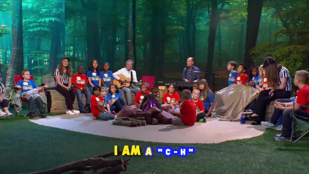 A group of children and adults are gathered around a fake campfire, while two men play music. The scene is set up like a forest, with a backdrop of trees.

