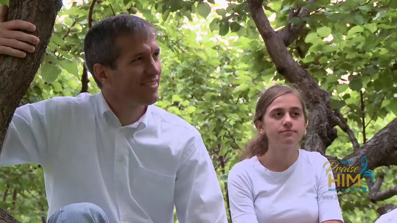 A man and a young woman sit in a tree, bathed in sunlight filtering through the leaves. The man, in a white shirt, looks off to the side, while the woman in a white t-shirt stares directly ahead.
