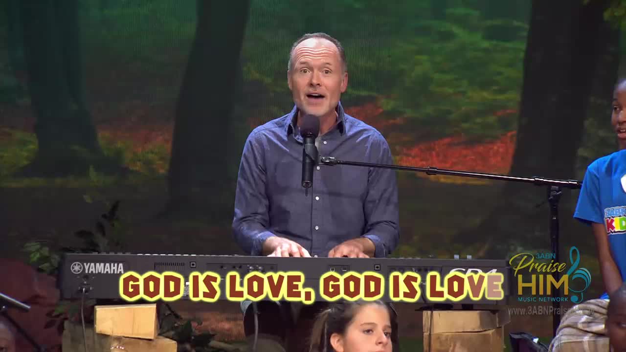 A man in a blue shirt plays a Yamaha keyboard, singing into a microphone. Children are visible on either side, and the words "GOD IS LOVE, GOD IS LOVE" flash across the screen during the 3ABN Praise Him Music Network broadcast.
