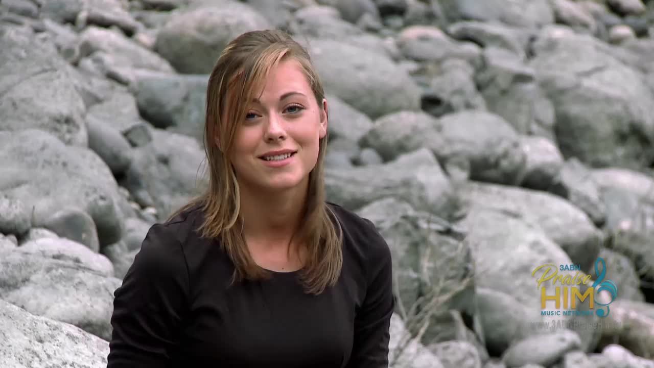 A young woman in a black shirt smiles as she speaks, the rocks behind her providing a neutral backdrop. The 3ABN Praise Him Music Network logo is visible in the corner.
