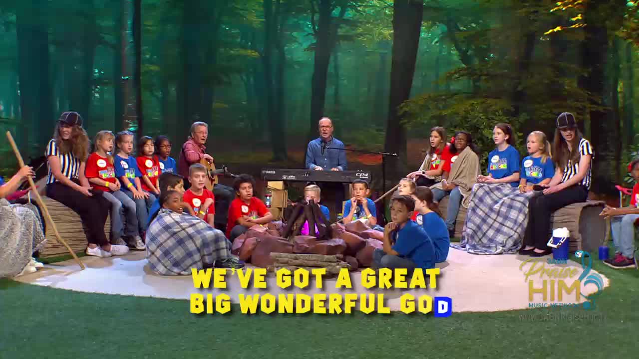 A group of children and adults are gathered around a fake campfire on a stage. The musicians in the center are playing, and the children are singing along to a song on the 3ABN Praise Him Music Network.
