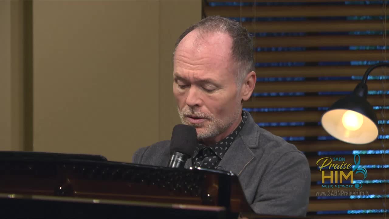 A man in a gray jacket sits at a piano, singing into a microphone. The 3ABN Praise Him Music Network logo glows in the corner.
