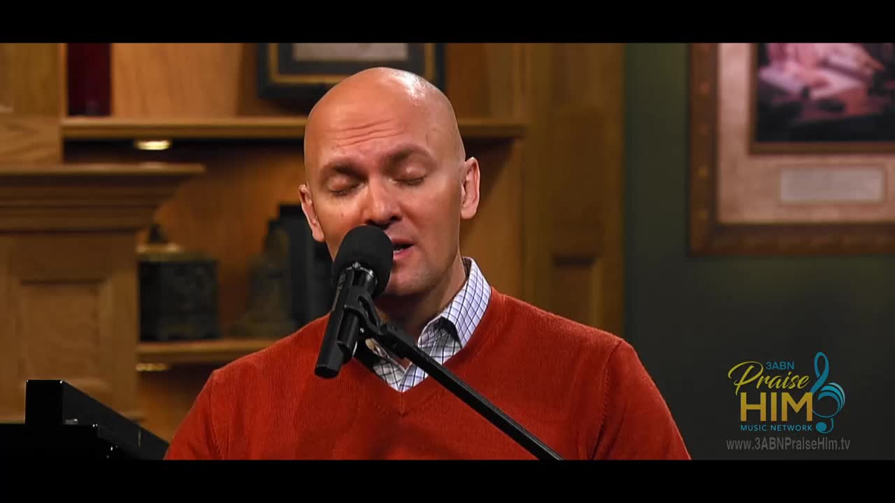 A man in a red sweater sings into a microphone, eyes closed. He's on a set, likely for a program on the 3ABN Praise Him Music Network.
