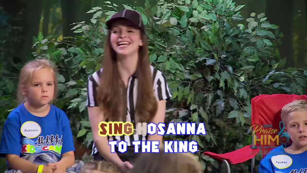 A young woman in a striped shirt and cap is smiling widely as she leads a group of children in a song. The words "Sing Hosanna to the King" are displayed on the screen, and the setting looks like a backyard, possibly for a program on the 3ABN Praise Him Music Network from the United States.
